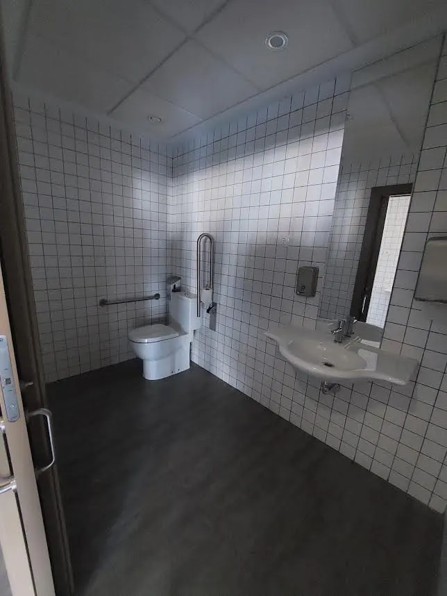 A bright, accessible bathroom with white tiled walls, a white toilet with grab bars, and a white sink. The floor is dark gray.