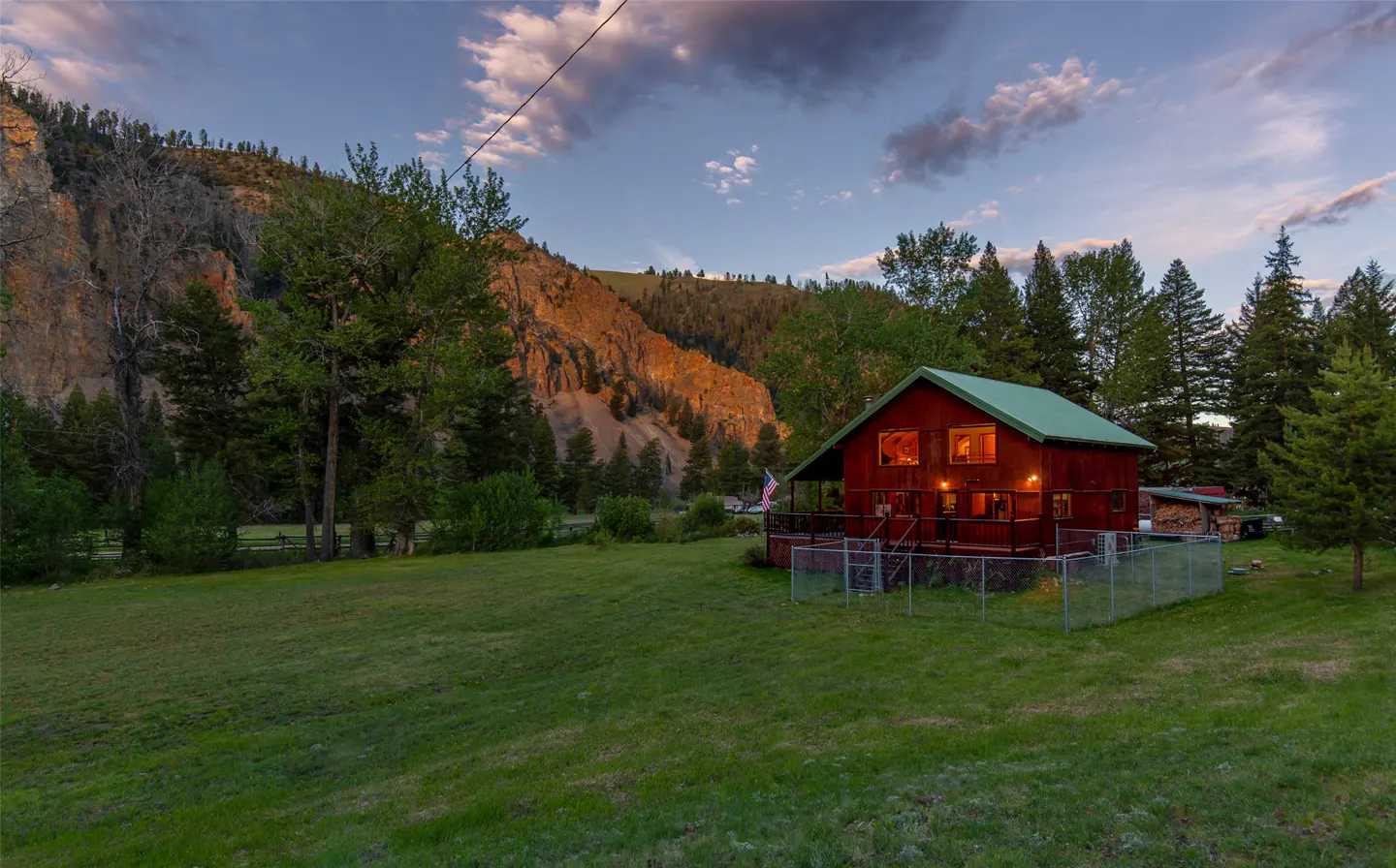 A red cabin with a green roof sits on a grassy lawn, backed by trees and a mountain at sunset.