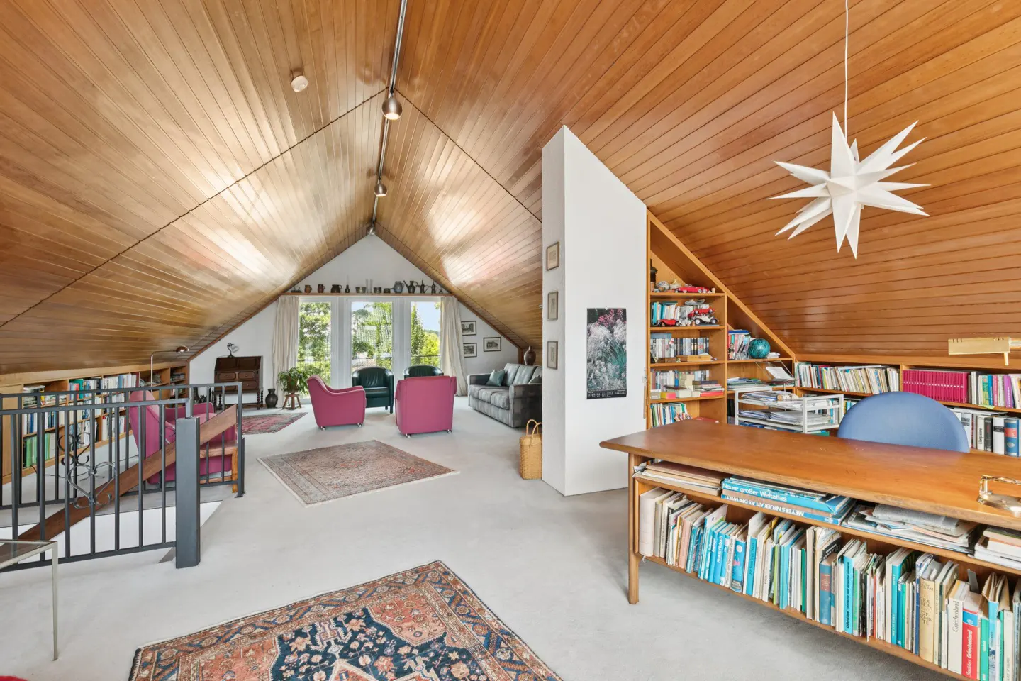 Attic living room with wood-paneled ceiling, bookshelves, desk, and seating area with pink chairs and gray sofa. A star-shaped lamp hangs from the ceiling.
