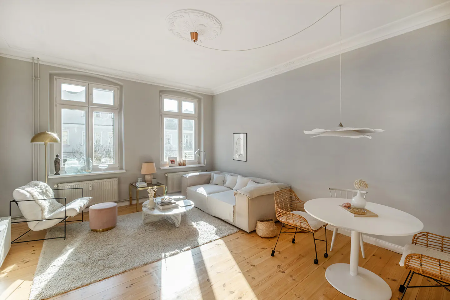 Bright living room with wood floors, gray walls, and white trim. A white sofa, chairs, and a round table create a cozy, modern space. Sunlight streams through the windows.