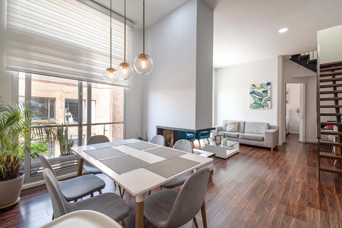 Bright, modern living space with a white dining table, gray chairs, and hardwood floors. Large window with blinds and hanging pendant lights.
