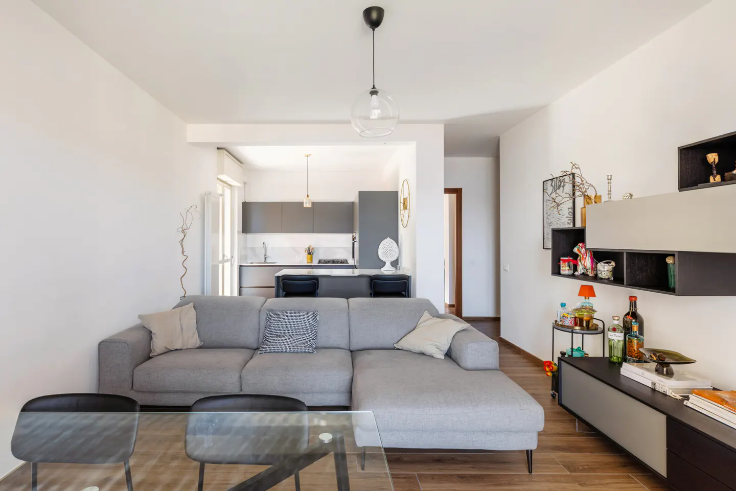 Bright living room with a gray sectional sofa, glass table, and open kitchen with gray cabinets. Wood floors and white walls create a modern space.