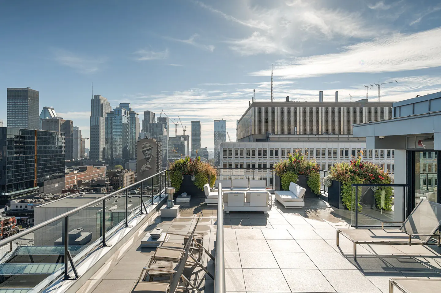 Rooftop terrace with white lounge furniture, potted plants, and sun loungers overlooking a sunny city skyline.