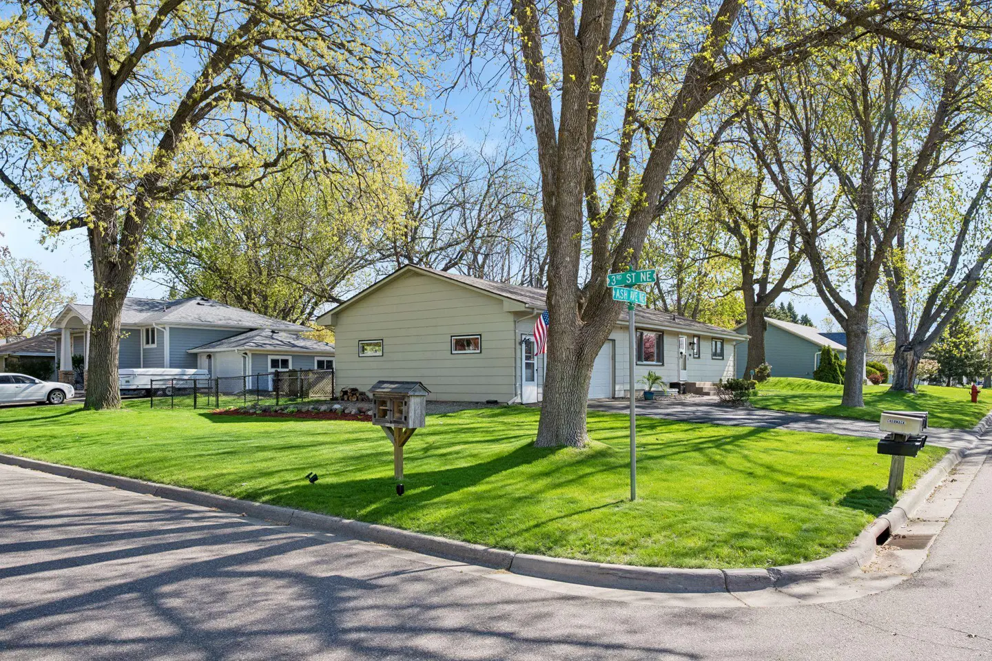 A single-story light green house on a corner lot with large trees and green grass. A street sign reads "3rd St NE".