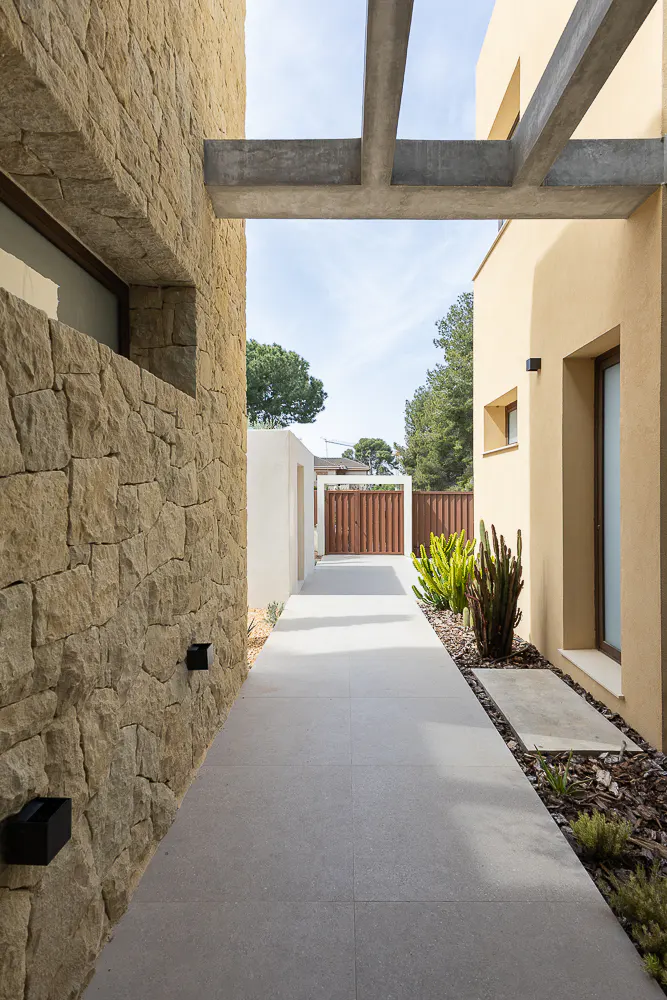 Exterior view of a modern home with stone and stucco walls, concrete beams, and a gray tiled walkway leading to a wooden gate.