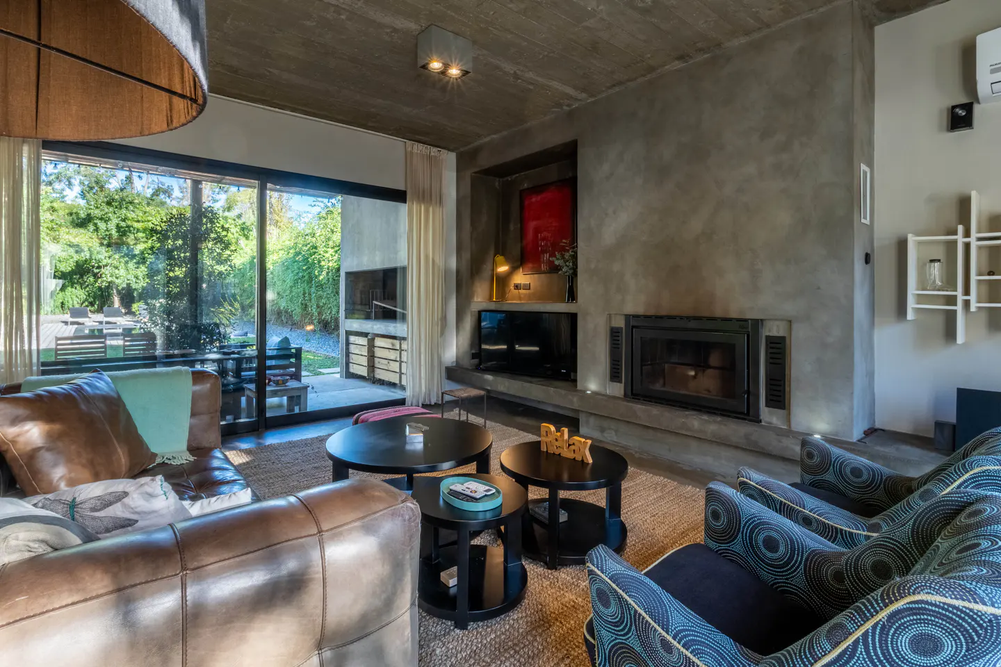 Living room with brown leather sofa, blue patterned chairs, black tables, and a fireplace. Large windows overlook a green backyard.