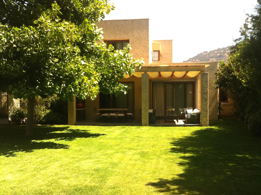 Tan stucco house with a covered patio, table, and chairs, surrounded by a green lawn and trees.
