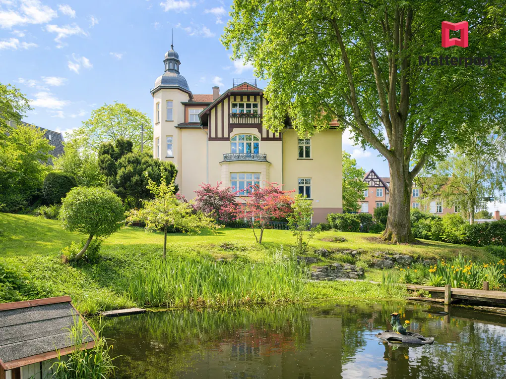 Exterior view of a large, light yellow house with a turret, surrounded by lush greenery and a pond reflecting the sky.