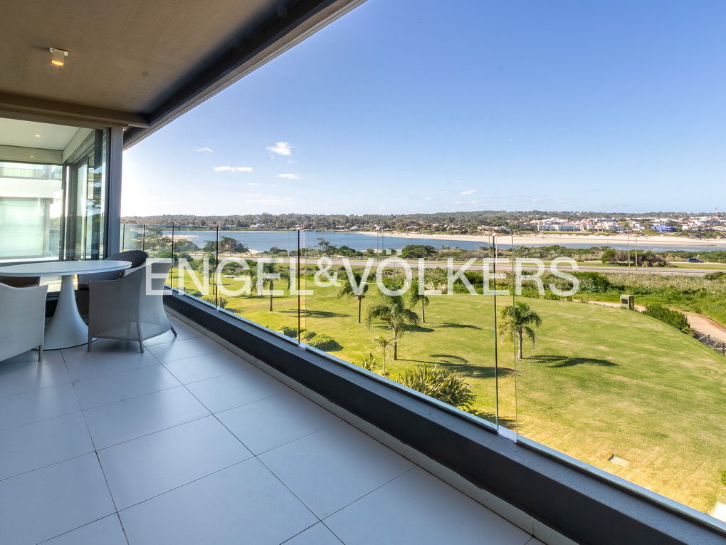 Balcony with white tile floor, table and chairs overlooking a green lawn, palm trees, and a body of water under a blue sky.