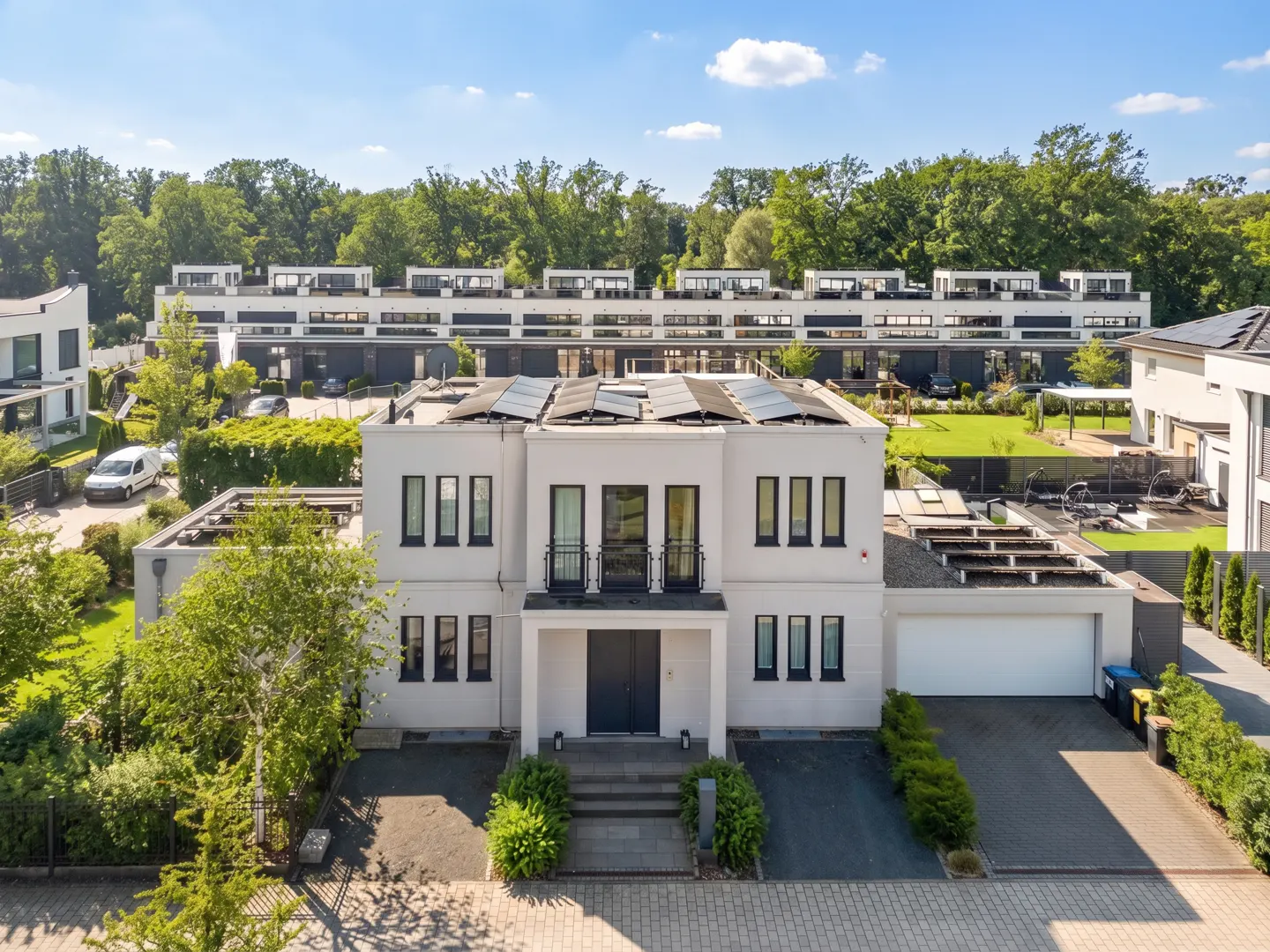 Aerial view of a modern, white two-story house with black trim, solar panels, and a two-car garage. Green trees in the background.