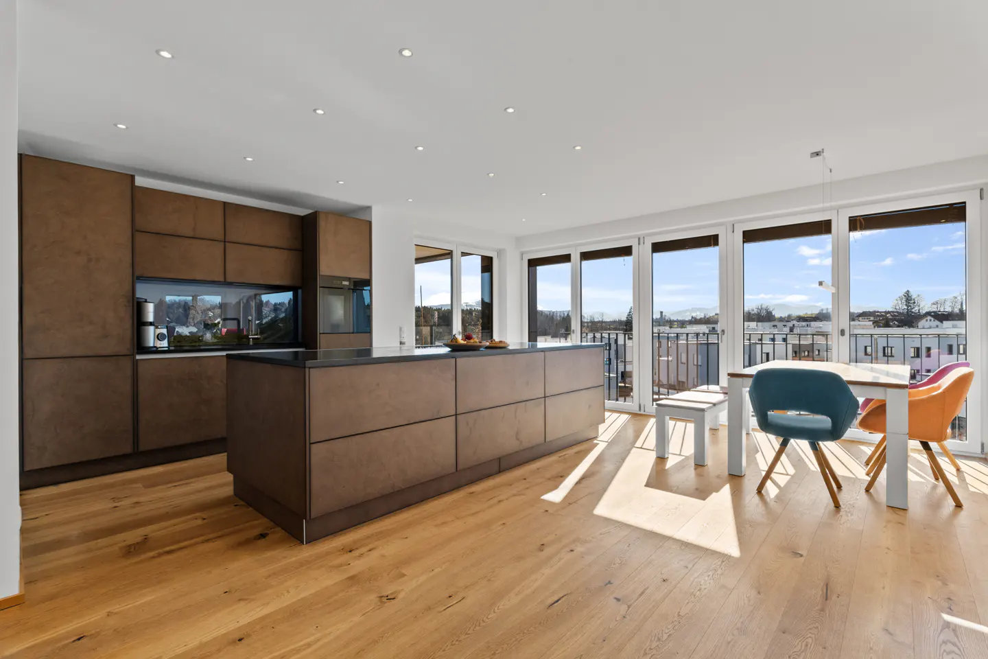 Bright, modern kitchen with brown cabinets, island, and wood floors. Dining table with blue and orange chairs near balcony doors.