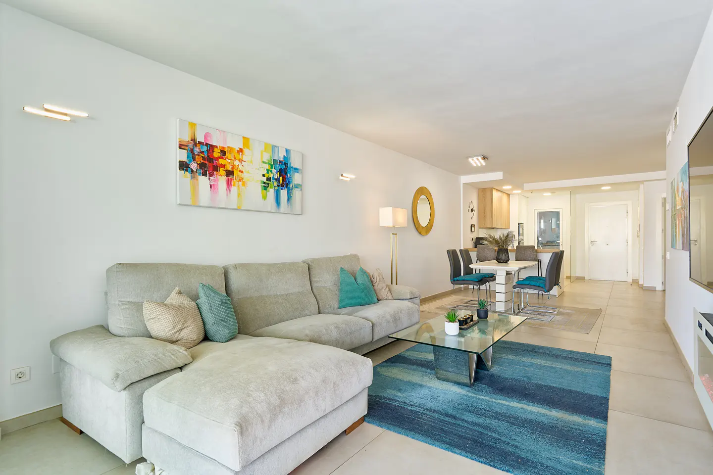 Bright living room with a gray sectional sofa, blue rug, glass coffee table, and dining area with white table and blue chairs. Colorful art on the wall.