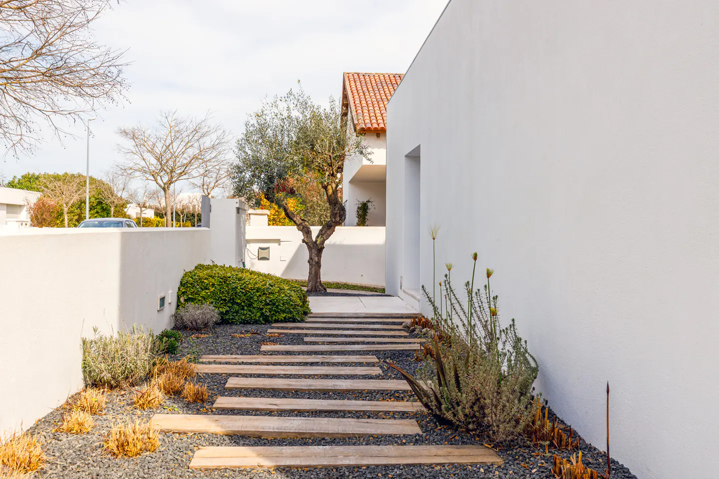 Modern home exterior with a walkway of wooden planks over black gravel, leading to a white building with a red tile roof. Green shrubs and a tree are visible.
