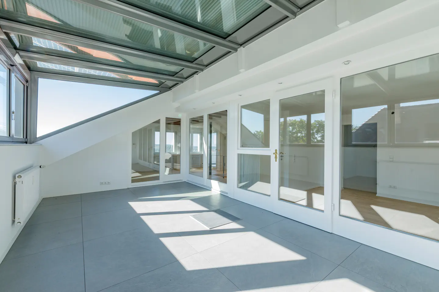 Bright sunroom with a glass ceiling, white walls, and gray tile floor. Windows and glass doors offer views of the outside.