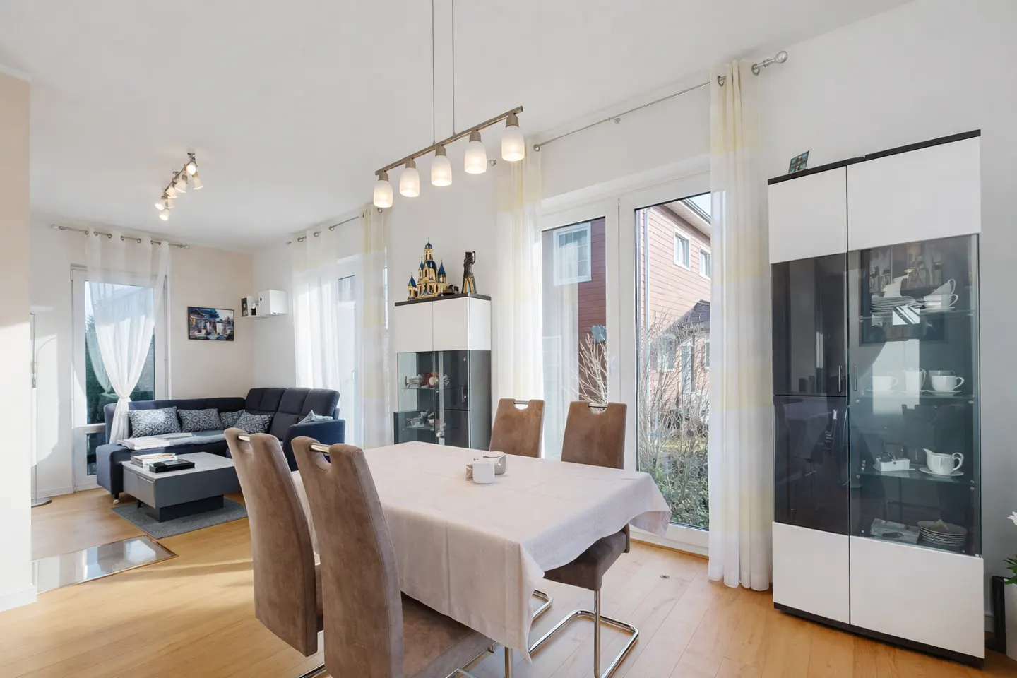 Bright, modern living room with a dining table set for four, a blue sofa, and a glass display cabinet. Light wood floors and white walls.