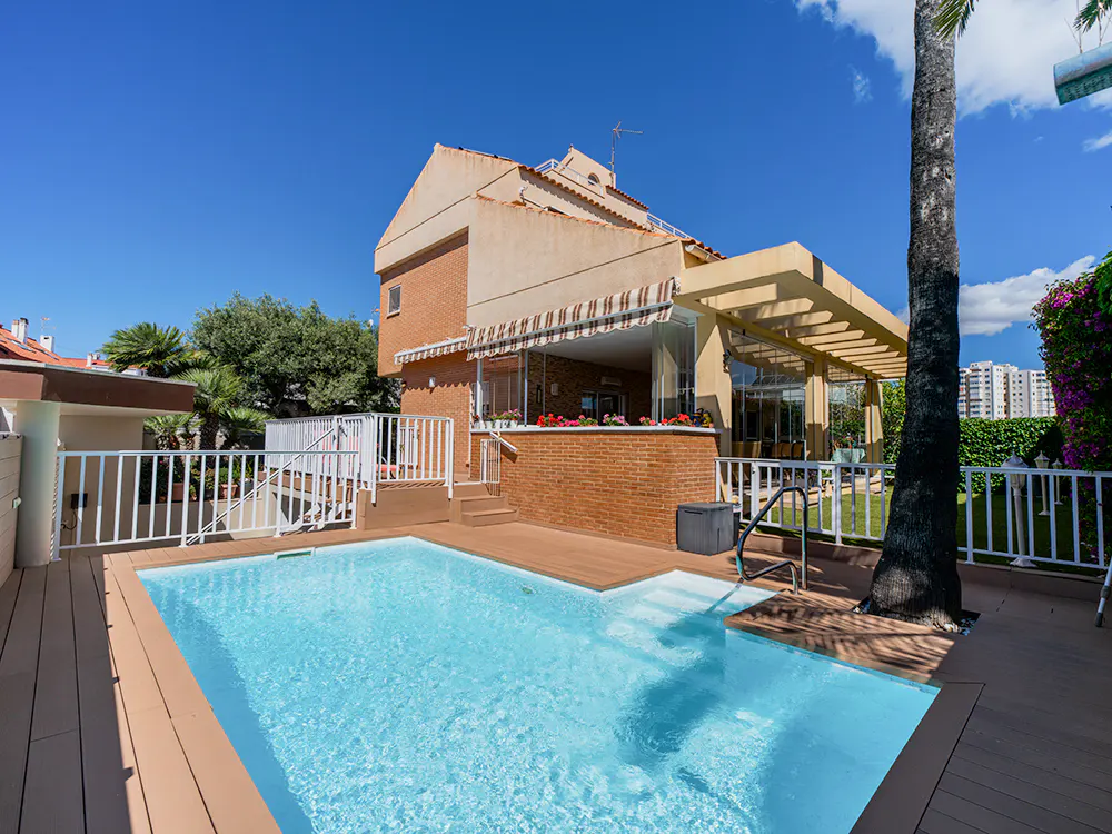 A backyard pool with clear blue water is surrounded by a wooden deck and a brick house under a bright blue sky.