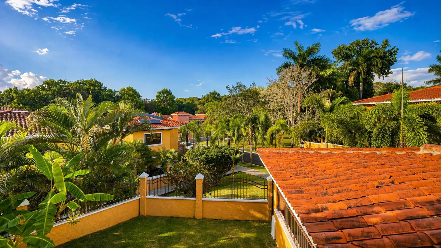 A sunny view of a neighborhood with yellow houses, orange tile roofs, and lush green trees.