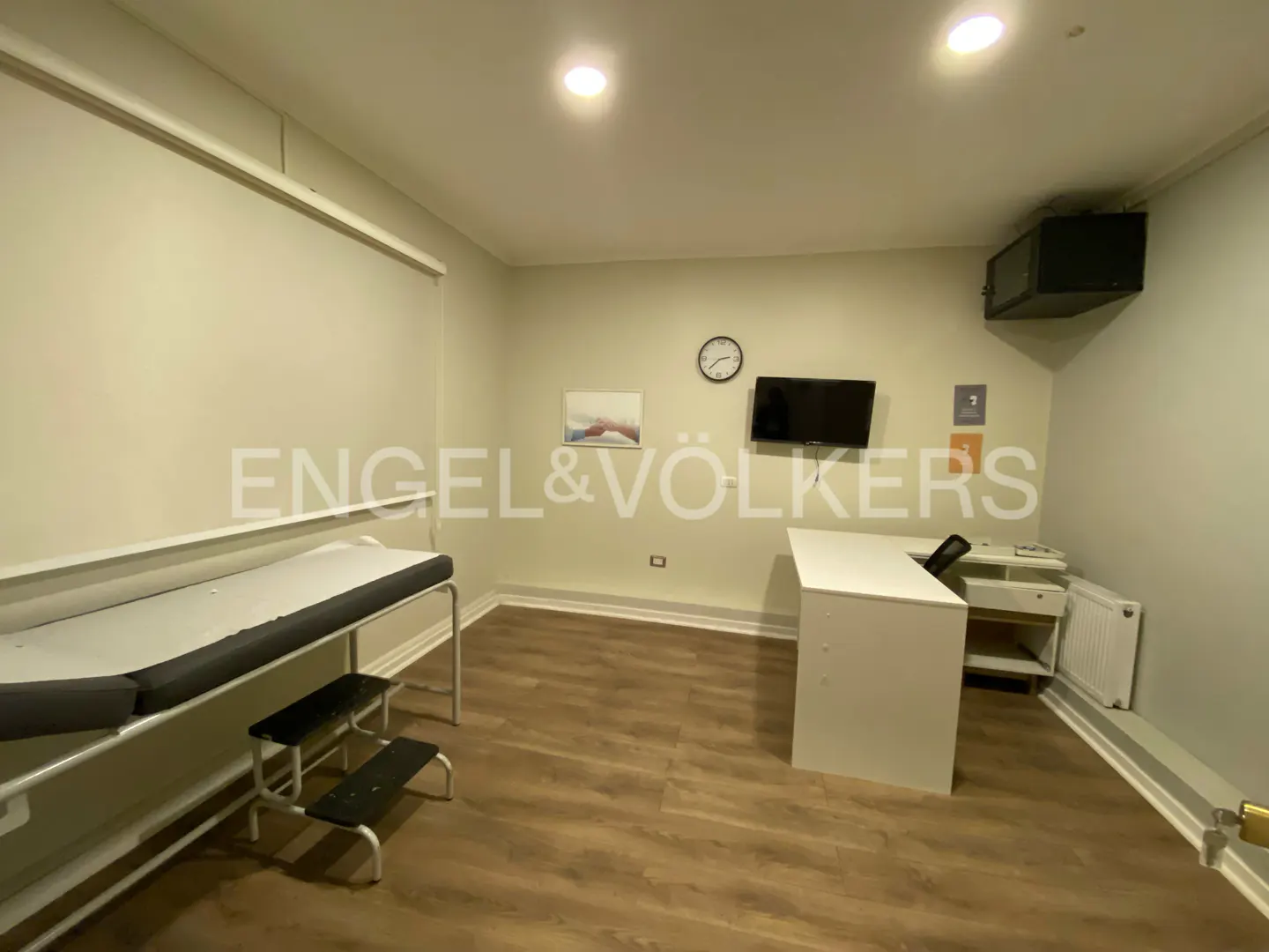 A doctor's office with an exam table, desk, and TV. The walls are light green and the floor is wood.