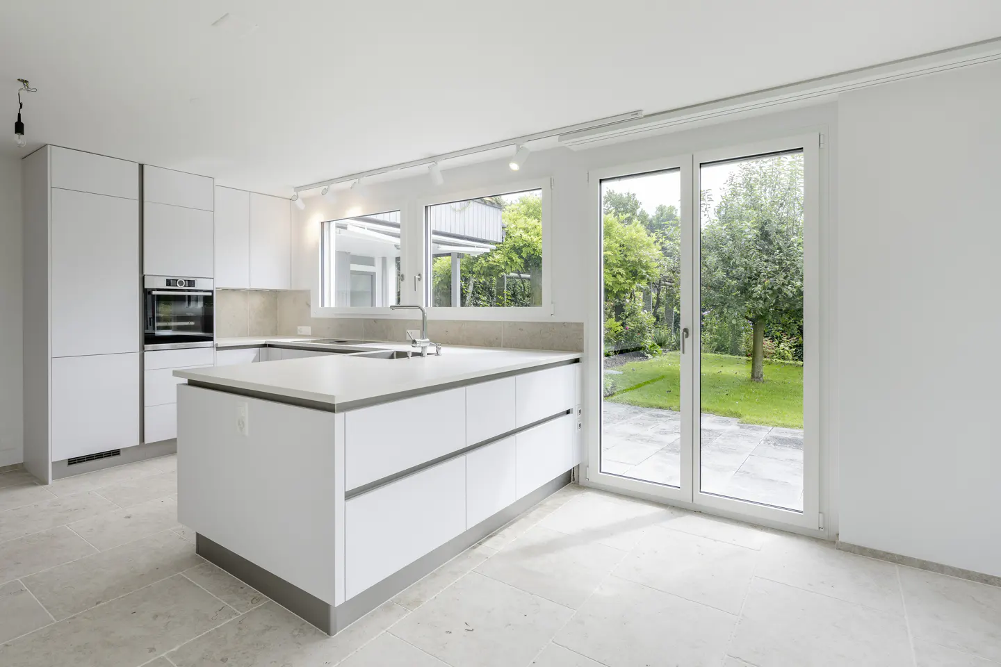 Bright, modern kitchen with white cabinets, island, and stone tile floor. Large windows and sliding glass doors offer a view of the green backyard.