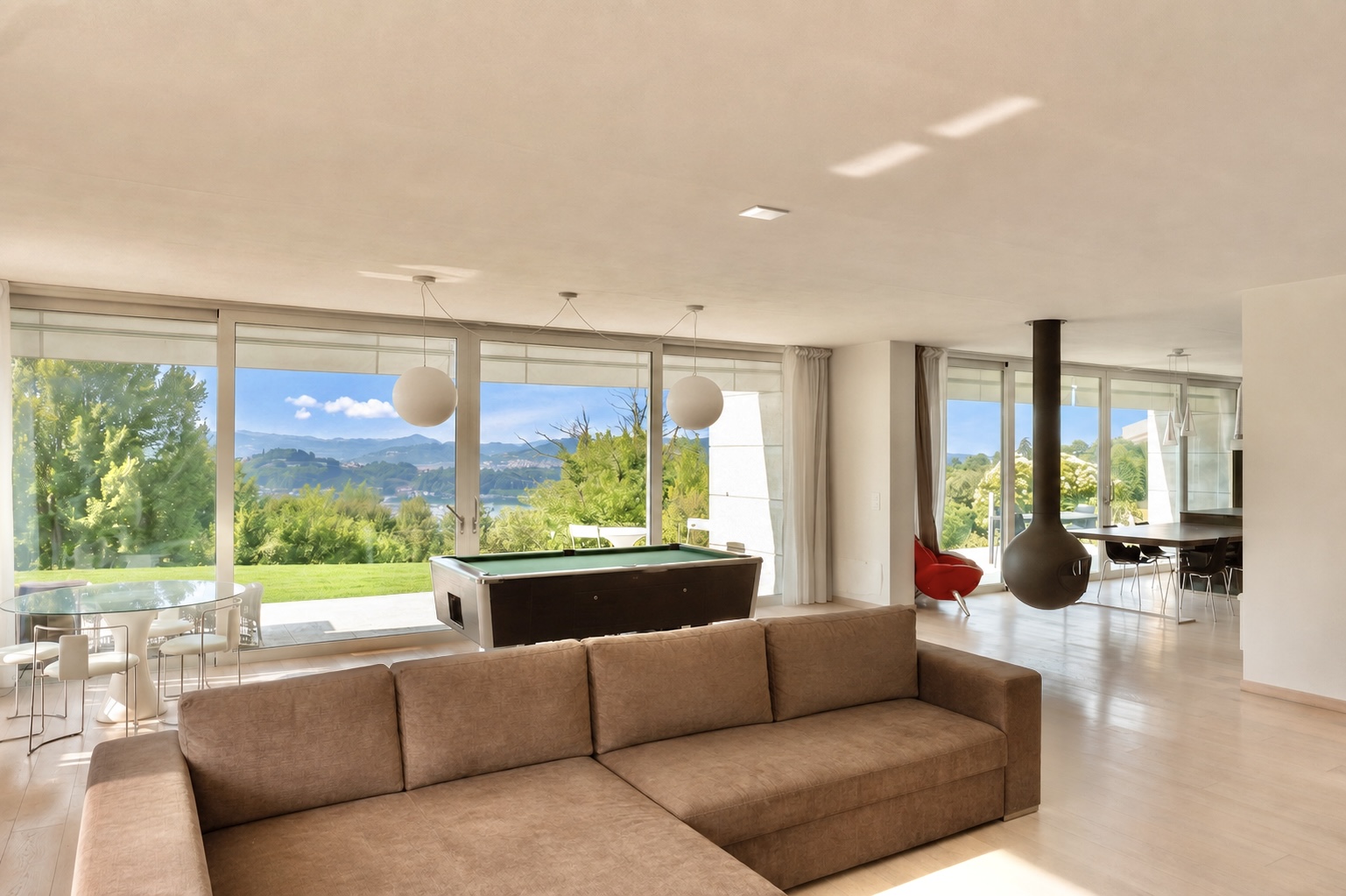Modern living room with a brown sectional sofa, pool table, hanging fireplace, and floor-to-ceiling windows with a view. [AI-generated text]