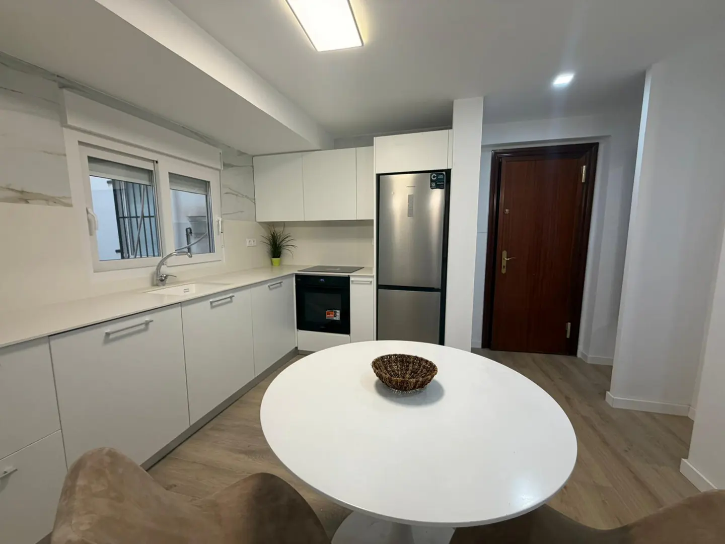 Bright kitchen with white cabinets, stainless steel fridge, and a round white table with a brown basket. A brown door is visible.