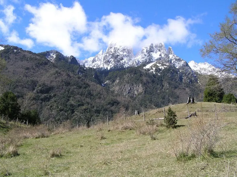 Scenic view of snow-capped mountains under a blue sky with white clouds, overlooking a green meadow with a wooden fence.