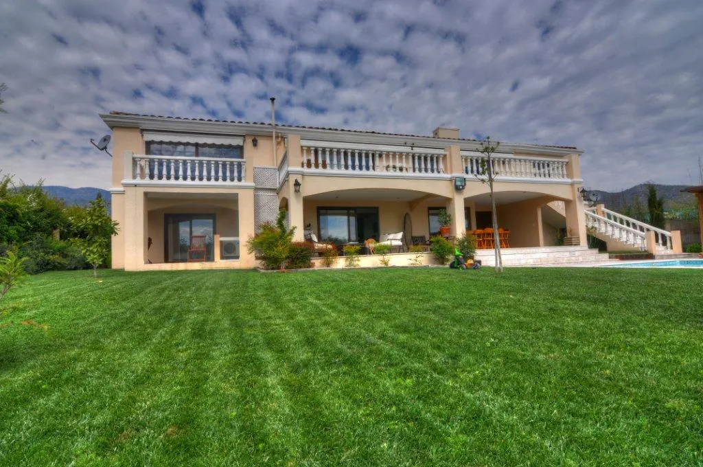 Two-story beige house with balconies overlooking a green lawn and pool. Cloudy sky.