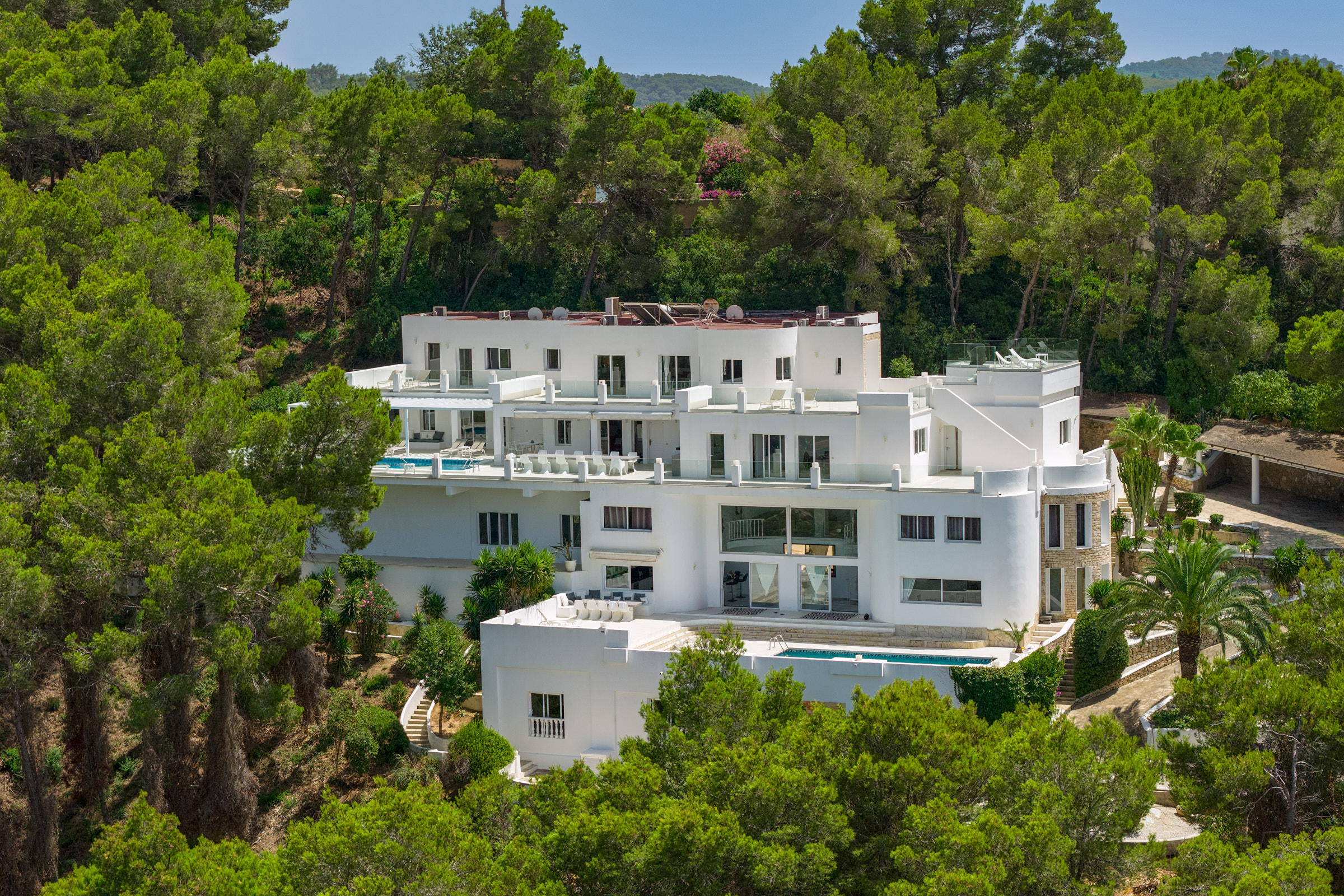 Large white multi-level villa with swimming pools and terraces, nestled in a lush green forest on a sunny day.