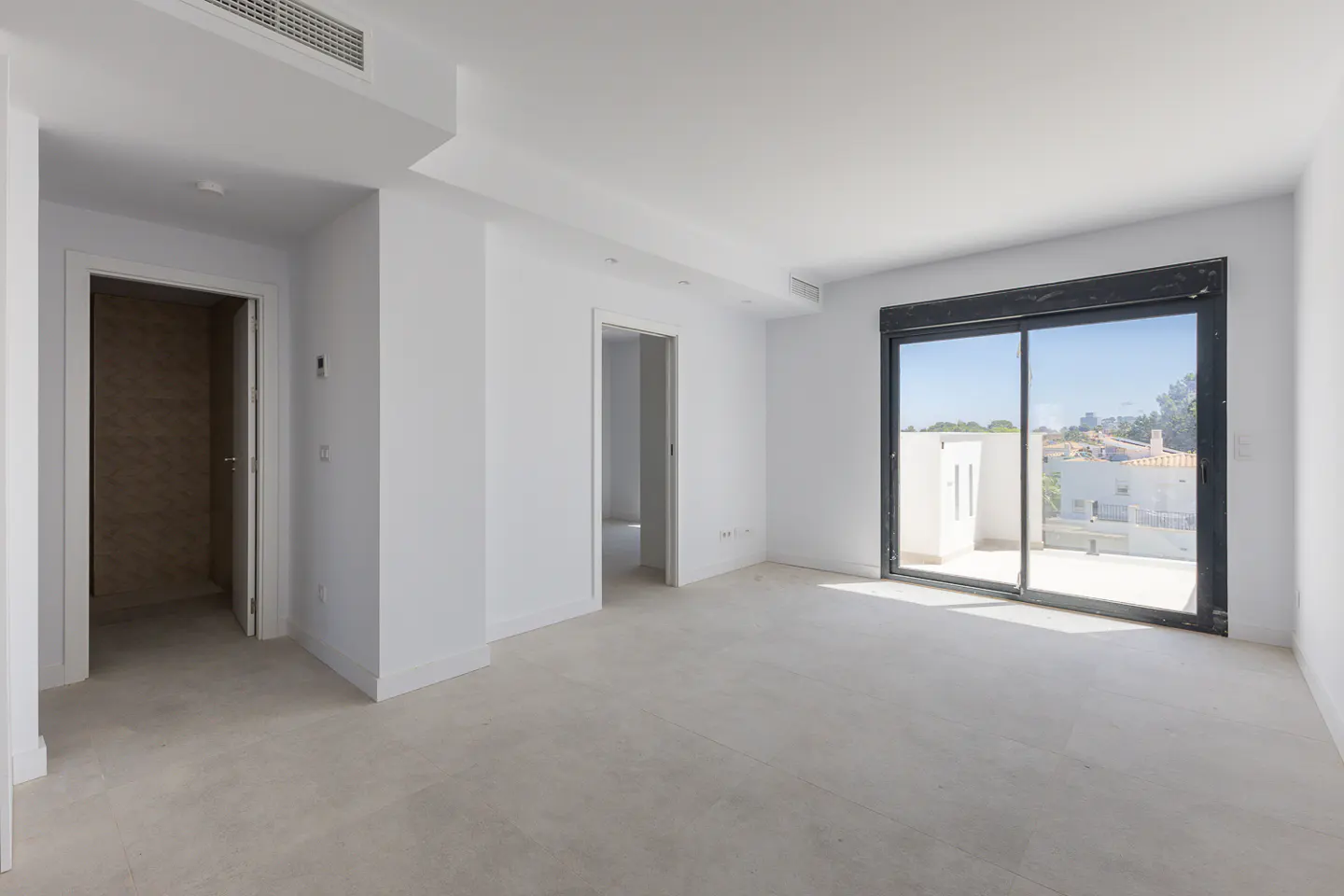 Empty, bright white room with light tile floors, two doorways, and a large sliding glass door leading to a sunny terrace.