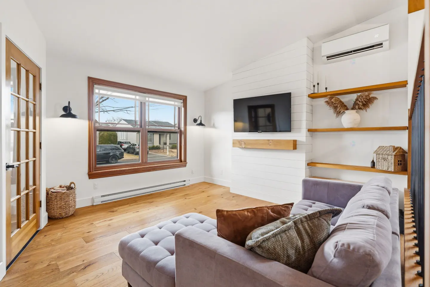 Bright living room with hardwood floors, a gray sectional sofa, and a window with a brown frame. A TV is mounted above a wooden shelf.