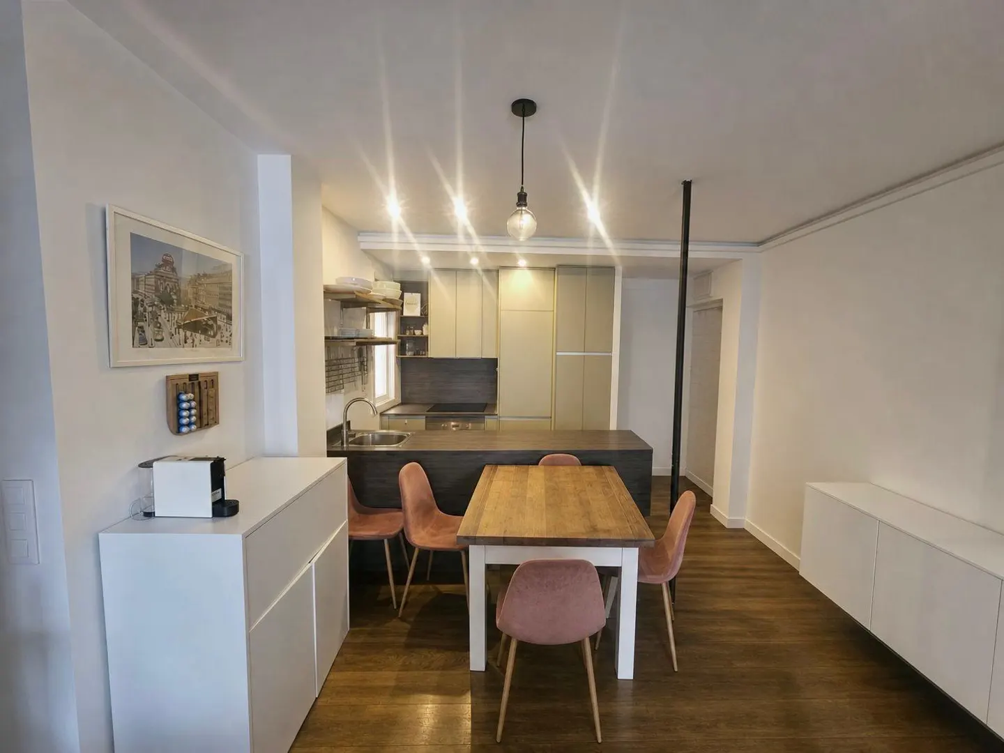 Bright, modern apartment interior with wood floors. A dining table with pink chairs sits near a kitchen with gray cabinets and a dark countertop. White cabinets line the walls.