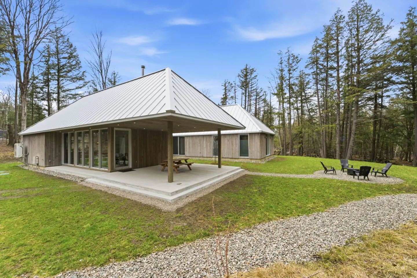 Modern cabin with a metal roof, wood siding, and a concrete patio with a picnic table, surrounded by green grass and tall trees.