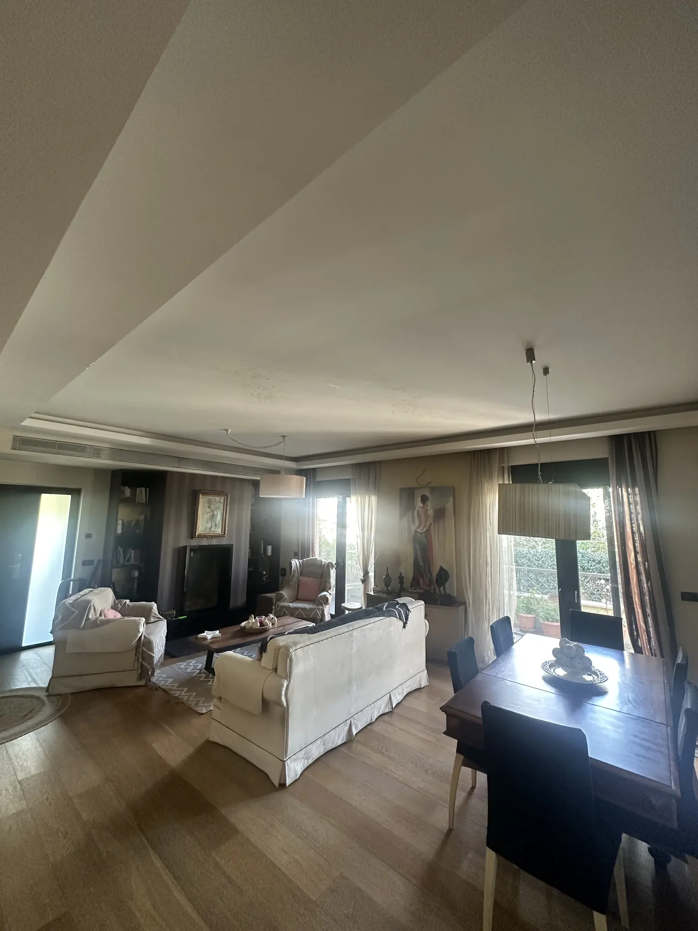 A bright, open-plan living and dining area with hardwood floors, a white sofa, and a dark wood dining table. Large windows offer views of greenery.