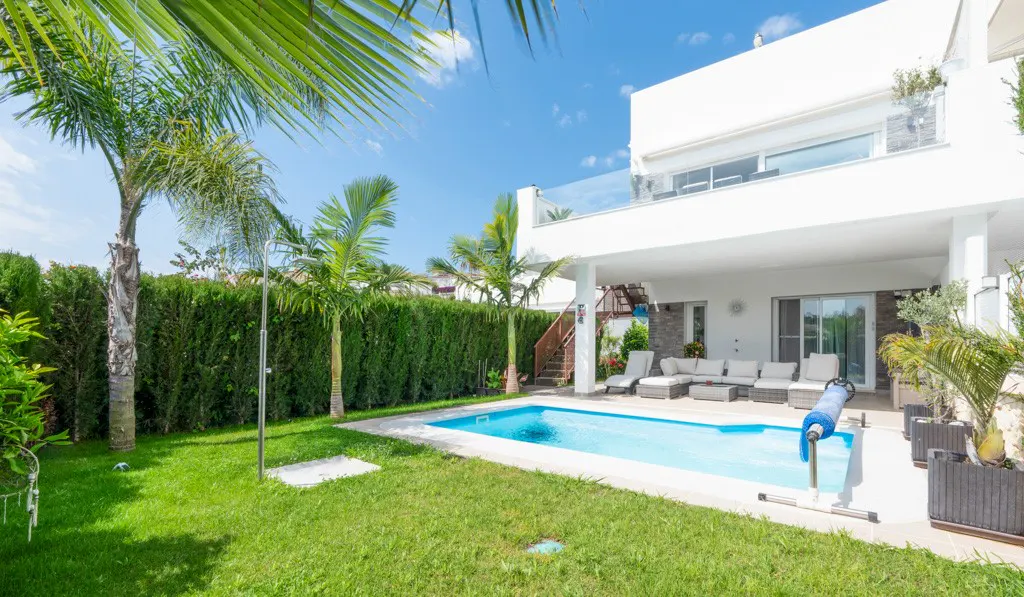 A modern white house with a pool, green lawn, and tall hedge under a blue sky with palm trees.