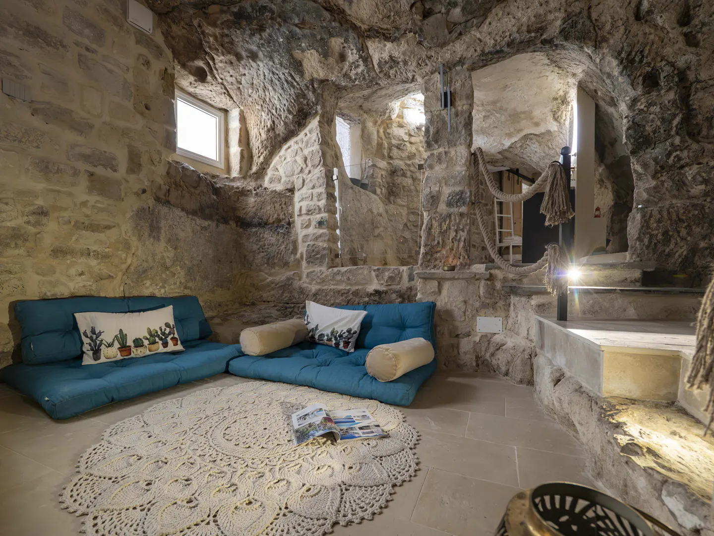 Cozy cave-style living room with natural stone walls, two teal floor sofas, a round crochet rug, and a rope railing.