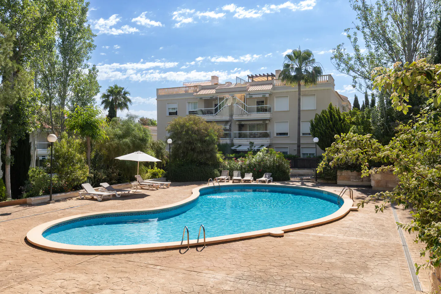 Kidney-shaped pool with lounge chairs and umbrella, set in a courtyard with a building and trees in the background.