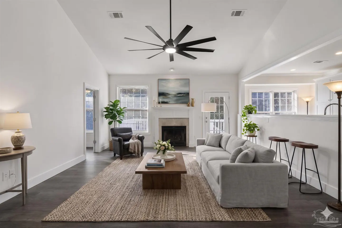 Bright, open living room with white walls, dark wood floors, and a large jute rug. A gray sofa faces a fireplace with an abstract painting above.