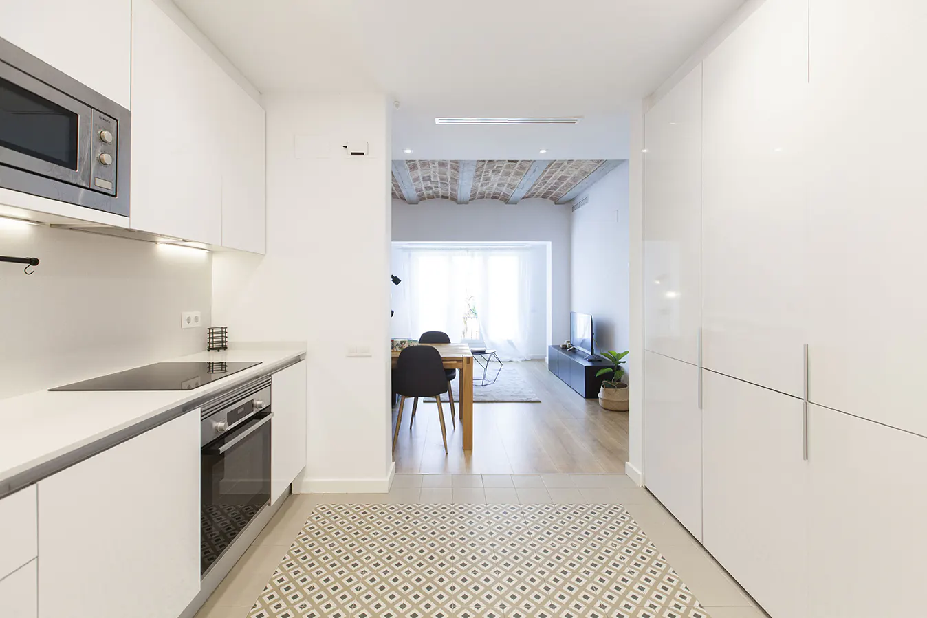 A bright, modern apartment interior. White kitchen cabinets line the left, leading to a dining and living area with a brick ceiling.