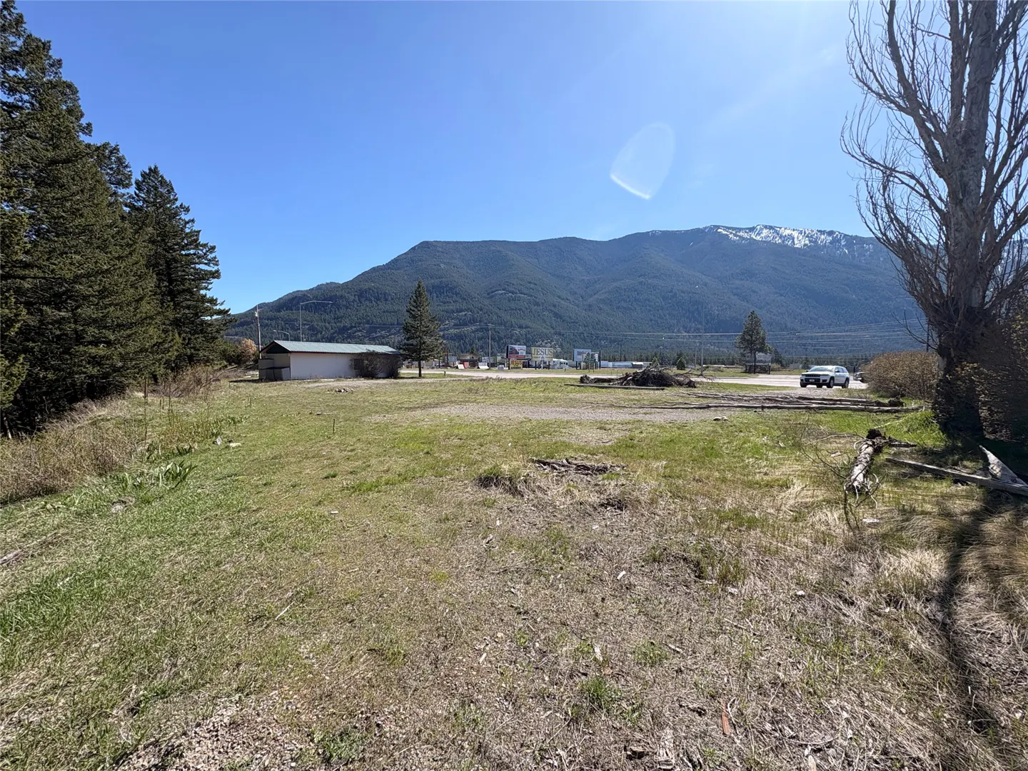 A grassy lot with a small building, trees, and a mountain backdrop under a clear blue sky.