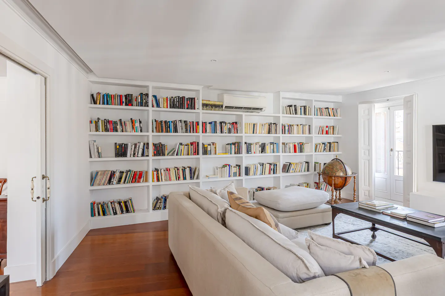 Bright living room with floor-to-ceiling white bookshelves filled with books. A beige sofa faces a dark coffee table with books. A globe stands near a window.