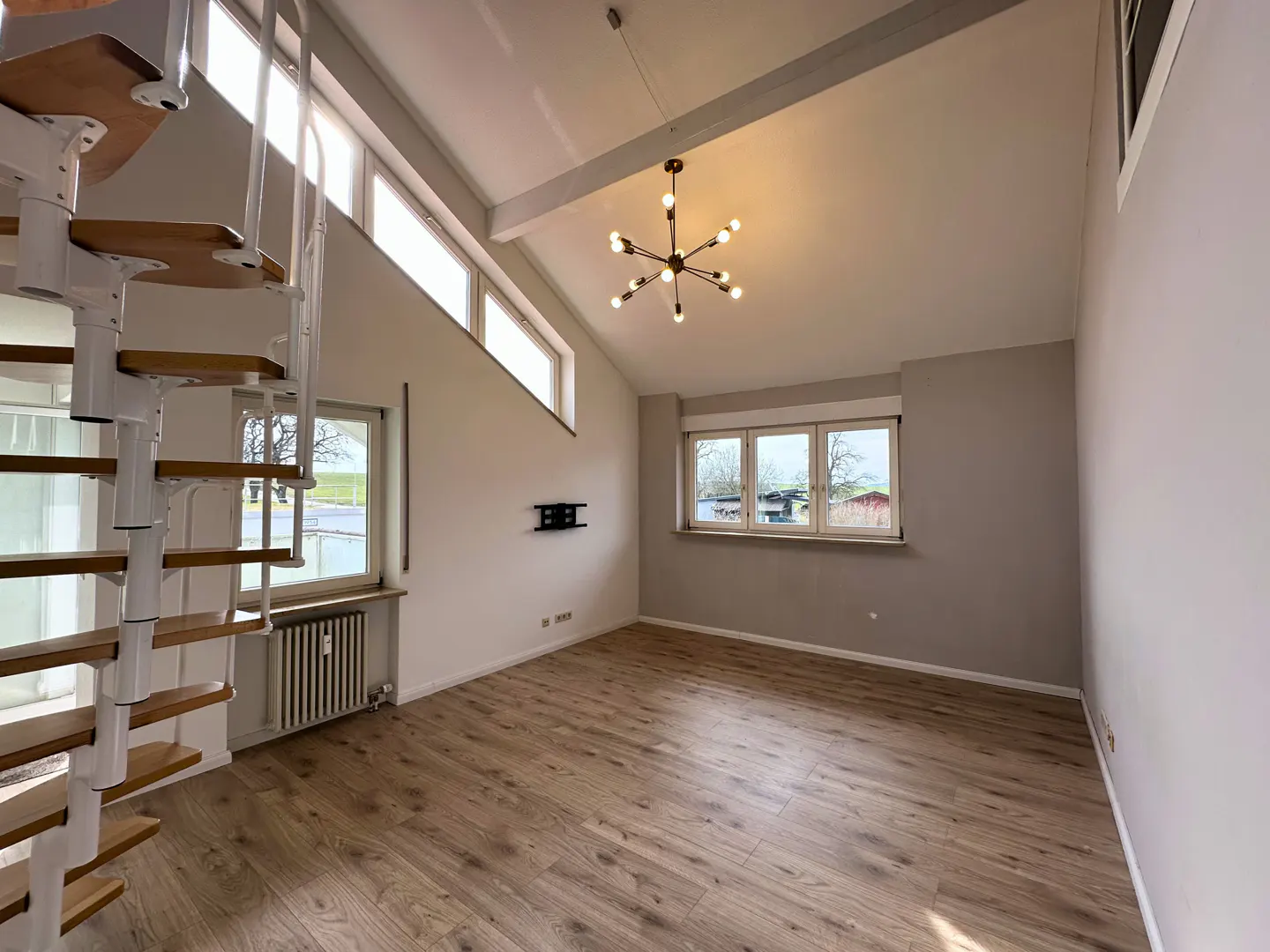 Bright, empty living room with wood floors, gray walls, and a white spiral staircase. A modern chandelier hangs from the vaulted ceiling. Windows offer outdoor views.