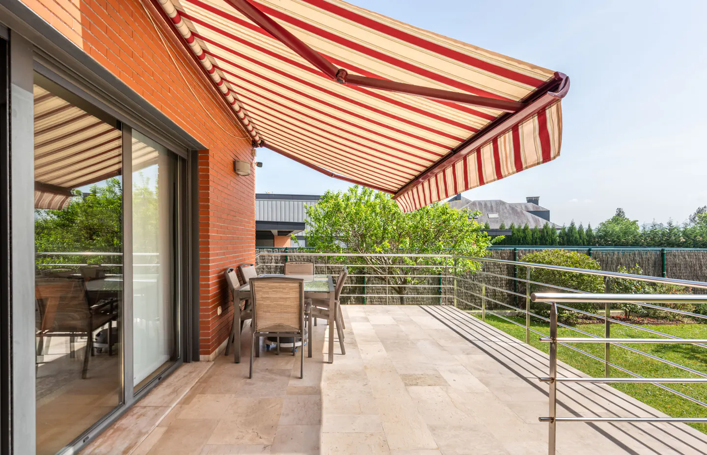 Outdoor patio with a red and white striped awning, table, chairs, and a metal railing overlooking a green lawn.