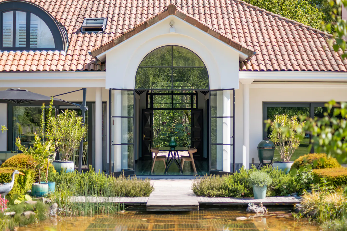 Exterior of a white house with a red tile roof, black framed arched doorway, and a pond in the foreground.