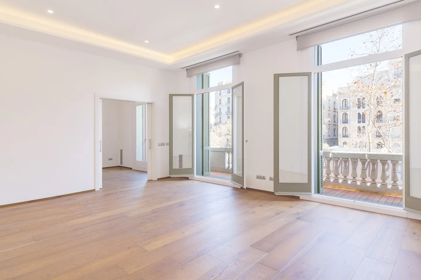 Bright, empty room with light wood floors, white walls, and open glass doors leading to a balcony overlooking city buildings.