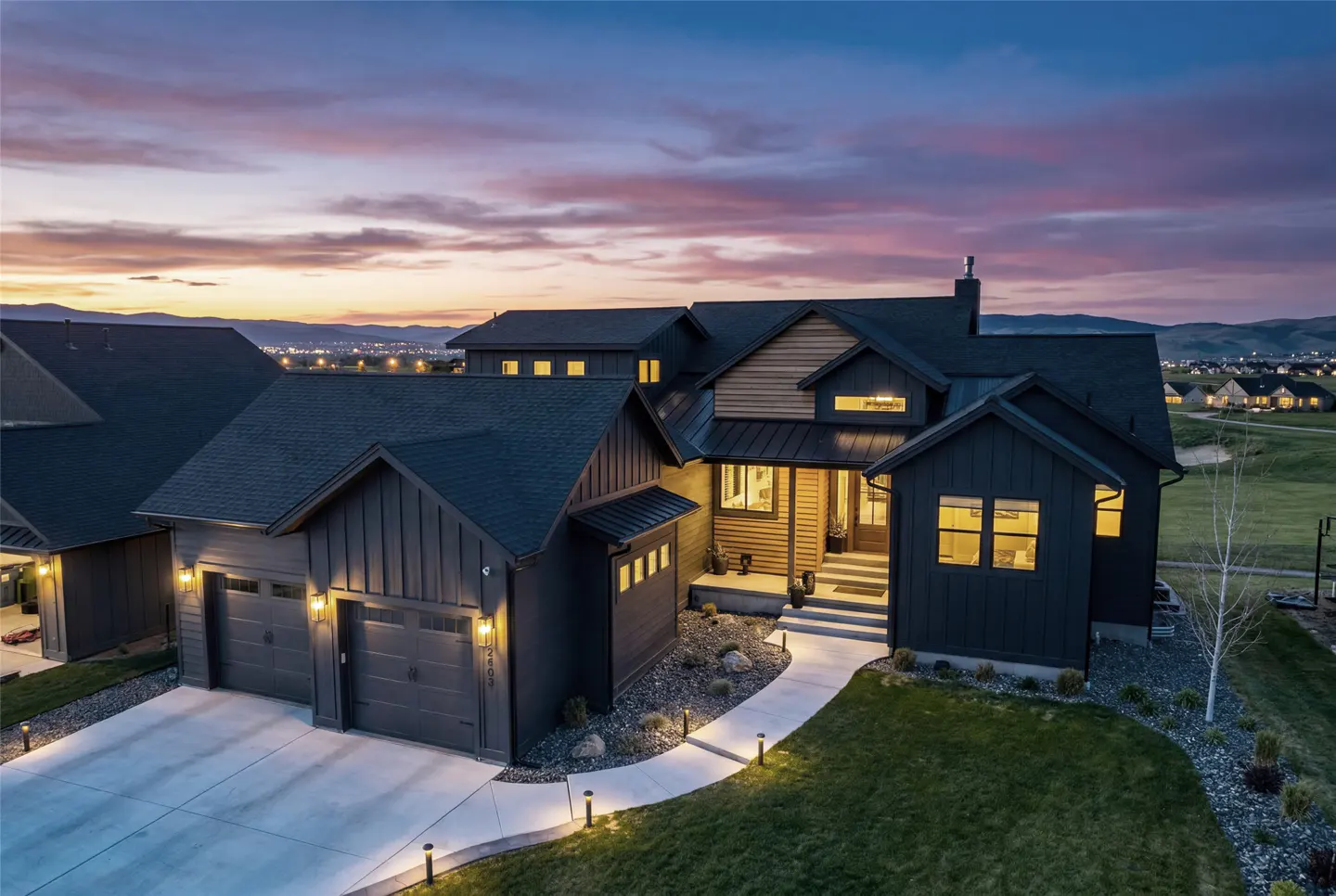 Aerial view of a modern, dark gray house with a two-car garage, a walkway, and a green lawn at dusk.