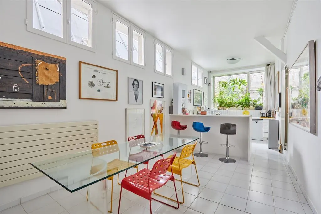 Bright, white dining room with a glass table, colorful chairs, and art-filled walls leading to a kitchen area with bar stools.