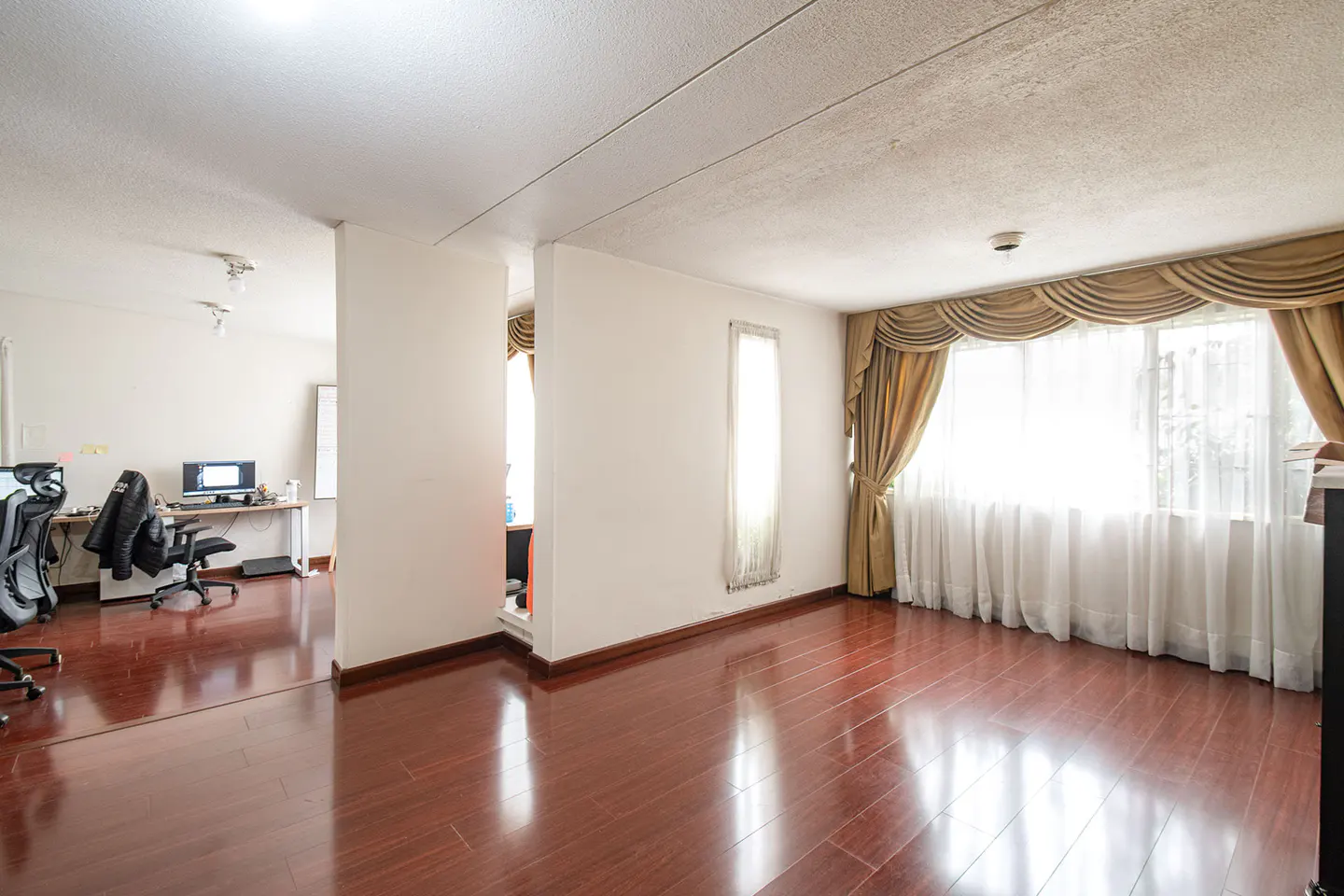 Bright room with hardwood floors, white walls, and gold curtains. An office space is visible through an opening.