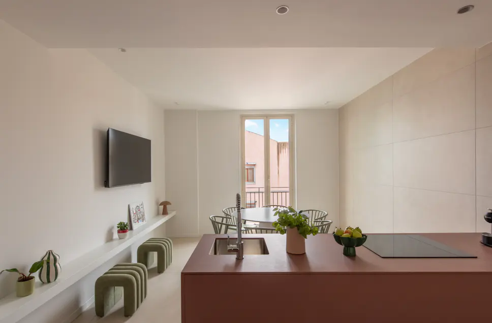 Bright, modern apartment interior with a brown kitchen island, a dining table near a window, and a TV on the white wall.