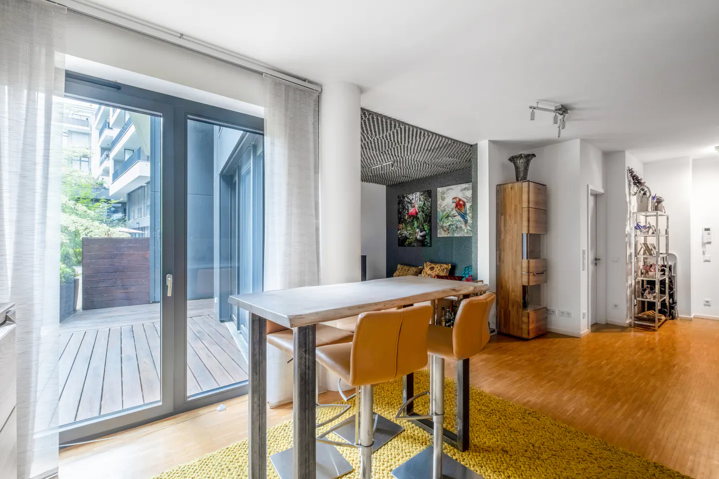 Modern dining area with a high table, two tan bar stools on a yellow rug, and a glass door leading to a wooden patio.