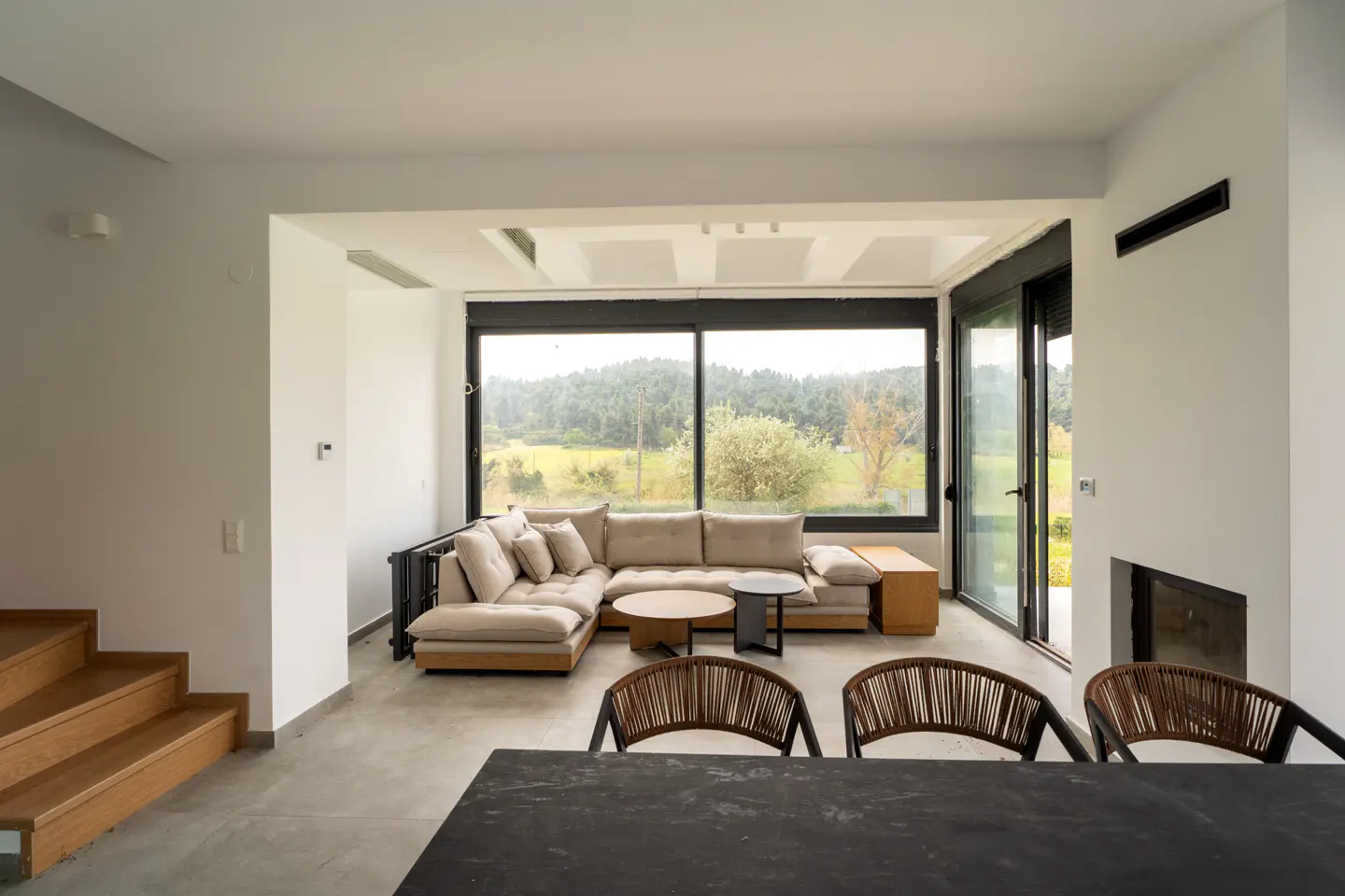 Bright living room with a beige sectional sofa, round tables, and woven chairs, with a large window overlooking a green landscape.